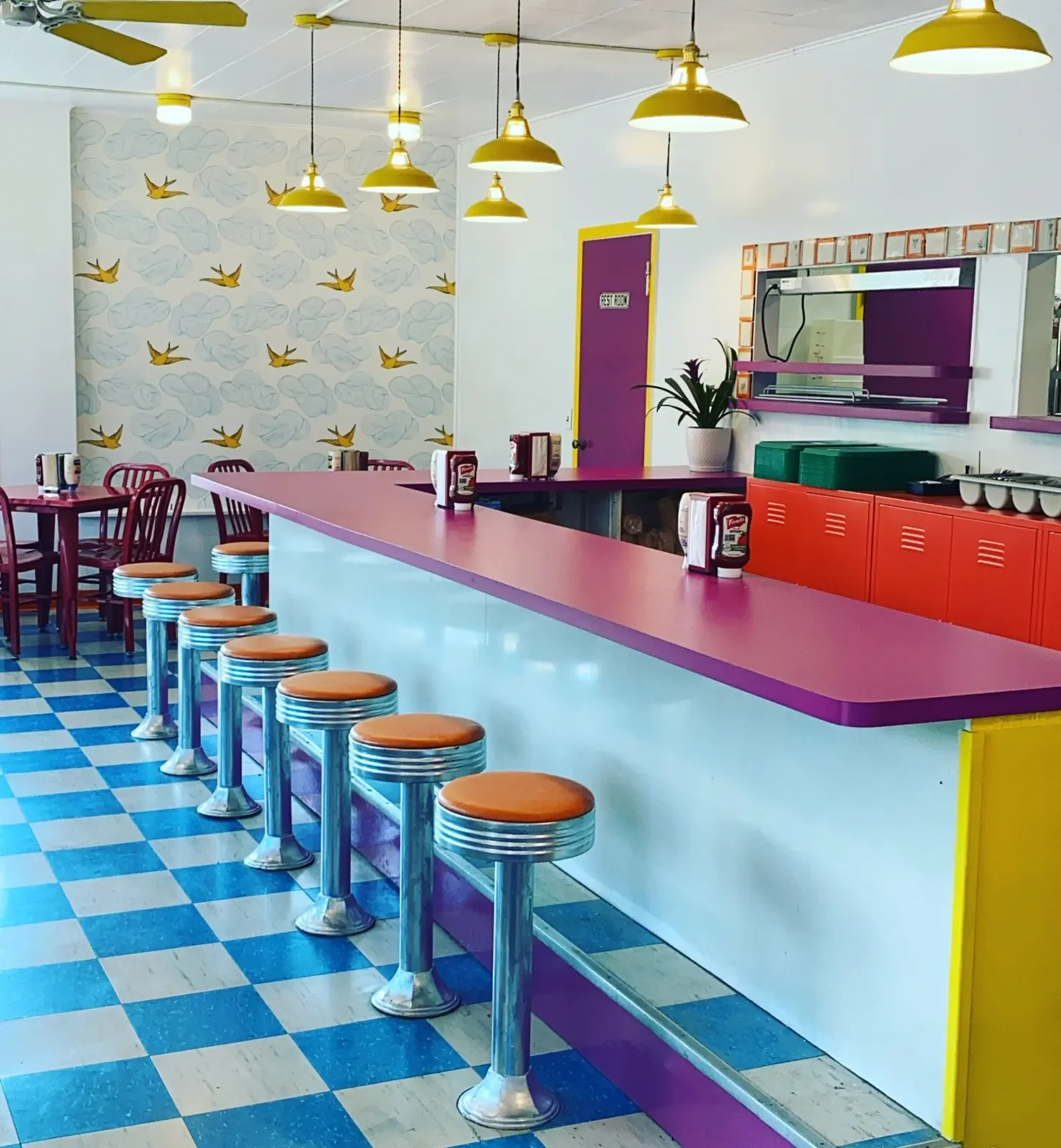 Inside the dinette: magenta counter, chrome stools with orange tops, blue and white checkerboard floor, mustard pendant lamps.
